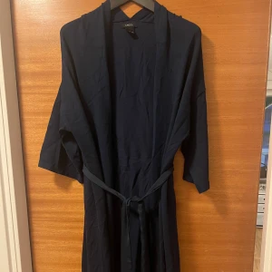 Mörkblå långkofta från Lindex - Mörkblå långkofta från Lindex med knytband i midjan och trekvartsärmar. Klänningen har en avslappnad passform och är tillverkad i mjuk bomull.