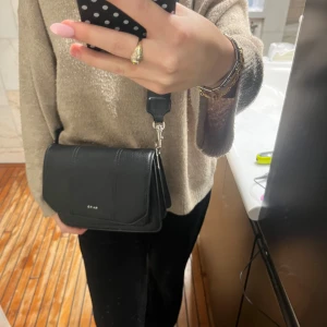 Svart axelväska från NA-KD - Snygg svart axelväska från NA-KD med bred axelrem och stilren design. Väskan är tillverkad i skinnimitation med struktur och har silverfärgade detaljer. Perfekt storlek för mobil, plånbok och andra essentials. Diskret logga framtill.