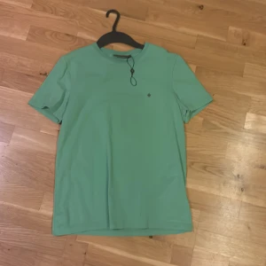 Grön t-shirt från Morris Stockholm - Fräsch grön t-shirt från Morris Stockholm i storlek XS. Klassisk rund hals och korta ärmar. T-shirten har en liten broderad logga framtill och är gjord i mjuk bomull som känns skön mot huden. Perfekt för dig som gillar stilrena plagg med diskreta detaljer. Helt ny lappen har kommit bort bara men bandet är kvar, helt oanvänd nypris 599