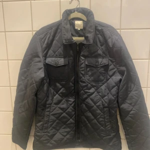 Mörkgrå quiltad jacka från Premium - Quiltad jacka i mörkgrått från Premium med två bröstfickor med lock och tryckknappar. Jackan har dragkedja framtill, klassisk krage och ett stilrent rutmönster. Perfekt för kyligare dagar under höst och vår.