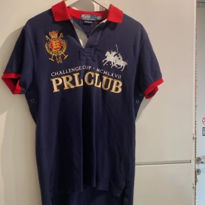 Blå Polo Ralph Lauren PRL Club piké - Snygg mörkblå pikétröja från Polo Ralph Lauren med röd krage och röda detaljer vid ärmslut. I princip oanvänd. Framsidan har broderat emblem i gult och rött, samt stor vit PRL CLUB-text och polospelare. Baksidan har tryckt PRL CLUB och siffran 3. Custom fit-modell i mjuk bomull.
