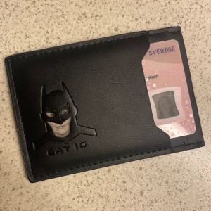 Svart Bat ID kortetui  - Slimmad svart plånbok med Batman-motiv och texten 'BAT ID' på framsidan. Plånboken har flera fack för kort och är tillverkad i mjukt fejkskinn. Perfekt för dig som gillar superhjältar och vill ha en stilren accessoar i fickan. Nytt.