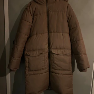 Beige lång pufferjacka med huva vinterjacka - Säljer en brun pufferjacka med huva och stora fickor framtill. Strl M. Från Object.