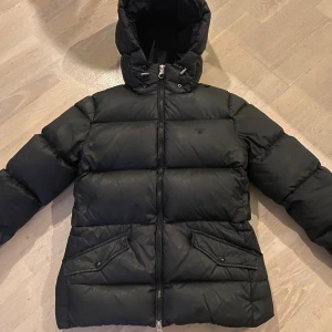 Svart dunjacka med huva från Emporio Armani - Svart dunjacka från Gant med huva och dragkedja framtill. Jackan har två stora fickor med lock och knappar, samt justerbar dragsko vid huvan. Puffig modell som håller dig varm under kalla dagar. Har täcken på användning men inget märkvärdigt, ordenarie pris runt 1500kr 💕