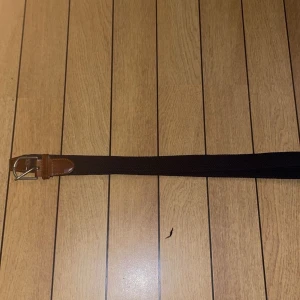 Svart flätat bälte med skinn-detaljer - Snyggt svart flätat bälte med bruna skinn-detaljer vid spännet och spetsen. Bältet har ett klassiskt silverfärgat metallspänne och passar perfekt till jeans eller chinos. Den flätade designen ger en modern och stilren look.