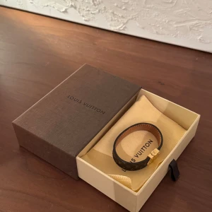 Louis Vuitton monogram armband guld - Snyggt armband från Louis Vuitton i klassiskt monogrammönster. Armbandet är brunt med guldigt spänne och detaljer, tillverkat i läder med ikoniska LV-loggor. Perfekt accessoar för dig som vill ha en lyxig och tidlös look.