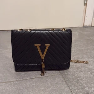 Svart quiltad axelväska med kedja - Snygg svart axelväska med quiltat mönster och guldfärgad kedja. Väskan har ett stort V i metall framtill med dekorativ tofs. Perfekt storlek för det viktigaste och ger en lyxig vibe till din outfit.