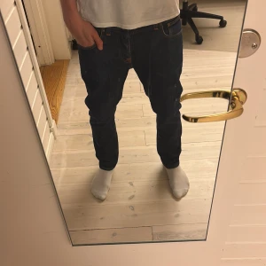 Mörkblå jeans från Nudie Jeans, W32 L32 - Säljer ett par snygga mörkblå jeans från Nudie Jeans i klassisk straight fit. Jeansen har orangea kontrastsömmar, fem fickor och normal passform. Materialet är slitstarkt denim i bomull och midjan är midwaist. Perfekt för dig som gillar stilrena och tidlösa jeans.