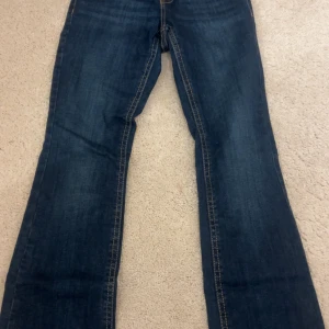 Blå bootcut jeans med fickdetaljer - Snygga blå jeans med bootcut passform och markerade sömmar. Baktill finns två fickor med lock och knappdetaljer, samt en läderpatch i midjan. Jeansen har klassisk femficksdesign och är tillverkade i slitstarkt denimtyg.