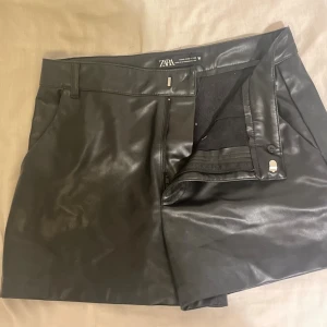 Svarta skinnshorts från Zara - Snygga svarta shorts från Zara i skinnimitation med  dragkedja fram. De har en clean look med fickor på sidorna och en stilren knappdetalj. Perfekta för dig som gillar edgy och trendiga plagg.✨😇