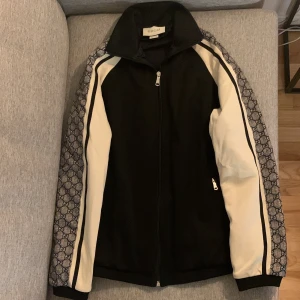 Svart och vit vindjacka från Gucci - Gucci vindjacka i svart med vita paneler och mönstrade blå/grå ränder längs ärmarna. Jackan har hög krage, hel dragkedja framtill och diskret logomönster på ärmarna. Snygg och sportig design som sticker ut med exklusiv känsla.