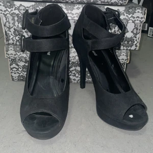 Svarta pumps med platå och spännen - (36, men passar på 38) Snygga svarta pumps från Graceland med hög klack och platåsula. Skorna har två breda remmar med stora spännen över vristen och öppen tå. Ytan är i mockaliknande material som ger en lyxig känsla. Perfekta för dig som vill sticka ut med en edgy look. Spännerna går även att stänga på 2 olika sätt. Rakt över, eller i ett X. 