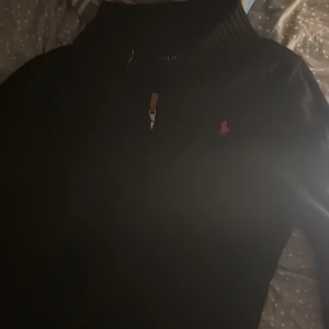 Svart stickad polotröja Polo Ralph Lauren - Svart stickad polotröja från Polo Ralph Lauren med half zip och ribbad krage. Klassisk röd logga broderad på bröstet. Perfekt för dig som gillar stilrena och tidlösa plagg med snygga detaljer.