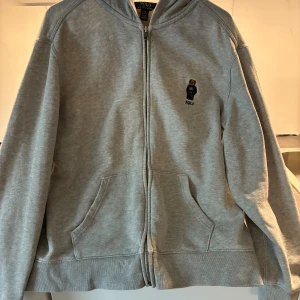 Grå hoodie från Polo Ralph Lauren - Grå hoodie från Polo Ralph Lauren med dragkedja och huva. På bröstet finns en broderad nalle med amerikansk tröja och texten POLO. Tröjan har två fickor framtill och är tillverkad i mjuk bomullsmix. Perfekt för chill dagar och streetstyle.