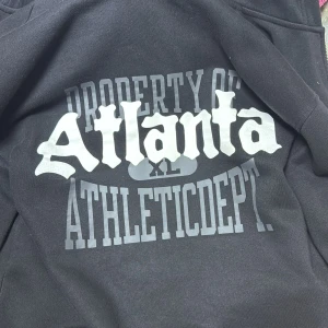 Svart hoodie Atlanta H&M M - Svart hoodie från H&M med tryck 'Atlanta Athletic Dept' i vitt och grått på bröstet. Relaxed fit med dragkedja framtill och mjukt material som känns skönt mot huden. Perfekt för chill dagar och streetstyle.