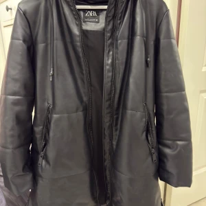 Svart puffer/skinnjacka med huva från Zara - Svart puffer/skinnjacka  från Zara med snyggt quiltad look och huva. Jackan har dragkedja framtill och två fickor med dragkedja på sidorna. Materialet är skinnimitation som ger en cool vibe och jackan har en loose passform. Perfekt för dig som gillar streetstyle.