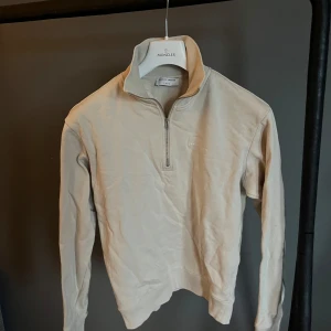 Tiger of sweden zip tröja  - Snygg beige långärmad tröja från Tiger of Sweden med half zip och hög krage. Tröjan är i mjuk bomull och har en stilren design med diskret logga på bröstet. Perfekt för dig som gillar minimalistisk och clean stil.