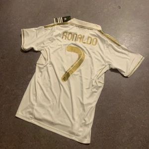 Real Madrid Ronaldo 7 Adidas tröja - Säljer en Real Madrid fotbollströja från Adidas med Ronaldo 7-tryck i guld på ryggen. Tröjan är vit med gulddetaljer, svart Bwin-logga på bröstet och klubbmärke. Tillverkad i lätt och ventilerande ClimaCool-material.