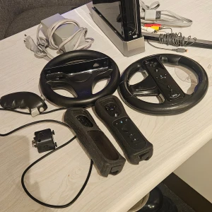 Nintendo Wii med tillbehör och spel - Komplett Nintendo Wii-paket med konsol, två rattar, två Wii-motes, nunchuk, Wii Fit Balance Board, strömadapter, kablar och 10 populära spel. Balance Board har viss missfärgning men allt är i fungerande skick. Perfekt för familjespel och träning! Säljer även spel o tillbehör separat om så önskas, finns i fler annonser. Kom med bud