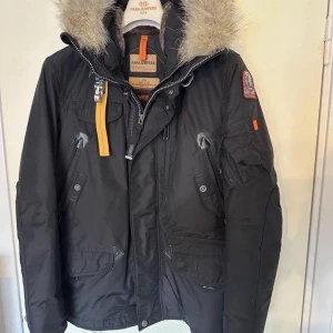 Parajumpers svart vinterjacka med päls - Svart Parajumpers Right hand vinterjacka parkas med snygg päls på huvan, ikonisk gul rem och patch på ärmen. Jackan har flera fickor, dragkedja och knappar, samt quiltat foder. Endast använd en säsong och i nyskick!