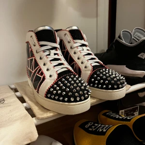 Christian Louboutin sneakers med nitar - Säljer ett par Christian Louboutin high-top sneakers i vitt, svart och rött med ikonisk röd sula. Skorna har coola silverfärgade nitar på tån, grafiskt mönster på sidorna och detaljer i skinn. Röd- och vitkantade sömmar och snörning framtill.