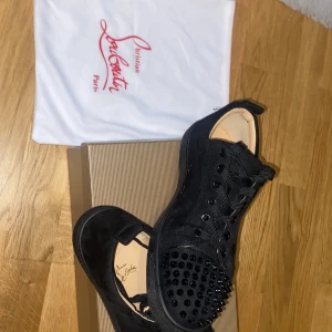 Christian Louboutin svarta sneakers med nitar - Svarta sneakers från Christian Louboutin i mocka med ikoniska röda sulor och svarta nitar på tån. Skorna har snörning och en platt sula, vilket ger en edgy och lyxig vibe. Perfekta för dig som vill sticka ut med exklusiva detaljer.