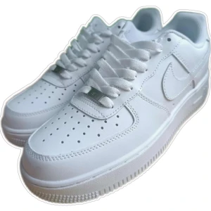 Nike Air Force 1 helvita sneakers - Klassiska Nike Air Force 1 sneakers i helvitt skinn med perforerad tå, vit sula och snörning. Snygg och tidlös modell med metallbricka på snörningen där det står AF1. Perfekta för dig som vill ha en clean och stilren look.