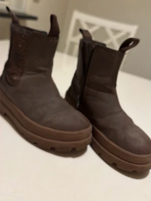 Bruna chunky chelsea boots - Säljer ett par bruna chelsea boots med grov platt sula och rund tå. Bootsens ovandel är i skinn och har elastiska paneler på sidorna för enkel på- och avtagning. Perfekt för dig som gillar robust stil och vill ha något som sticker ut.