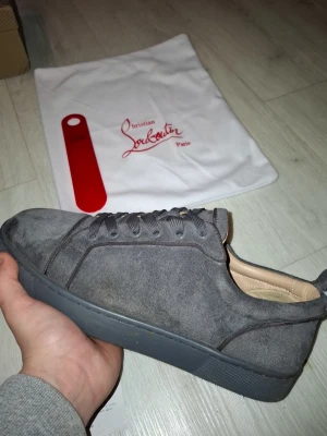 Grå mocka sneakers från Christian Louboutin - Säljer ett par gråa sneakers från Christian Louboutin i mjuk mocka med klassisk röd sula och rund tå. Skorna har snörning och platt sula, samt beige innersida. Kommer med originalkartong, dustbag och skohorn. Perfekt för dig som gillar exklusiva märken och stilren design.