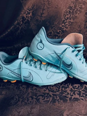 Nike blå fotbollsskor med snörning storlek 35 ( 22cm) - Säljer ett par blå Nike fotbollsskor med orange dobbar och snygga grafiska detaljer. Skorna har snörning och är tillverkade i syntetmaterial med meshfoder för extra komfort. Perfekta för dig som vill sticka ut på planen med stilrena och sportiga skor.