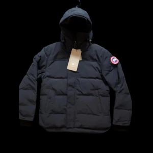 Canada Goose Winter Puffer Jacket – Hooded – Dark Navy – Unisex - Säljer en Canada Goose vinterjacka i puffer-modell i klassisk mörkblå färg. Jackan har tjock och varm vaddering, fast huva samt det ikoniska Canada Goose Arctic Program-märket på ärmen.   Perfekt för kalla vinterdagar, vardagsbruk, stadsmiljö eller utomhusaktiviteter. Tidlös och stilren design som passar både casual och mer uppklädda outfits.  Skick: Mycket bra / som ny (se bilder) Passform: Normal till något liten i storlek, L passar M och S.  Säsong: Höst / Vinter Stil: Lyx, vinterklassiker.