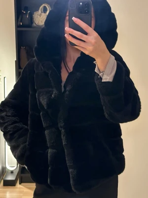 Svart pälsjacka - Svart pälsjacka med luva, knappar som stängning och fickor. Använd gåtal gånger och är i nyskick.