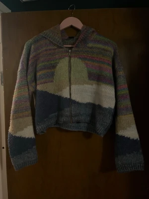 Flerfärgad stickad hoodie med dragkedja - Unik stickad hoodie med dragkedja och huva. Tröjan har ett färgglatt mönster i blått, grönt, beige, vitt och lila, samt inslag av randiga partier. Perfekt för dig som gillar mysiga och kreativa plagg med lite retrovibe. 