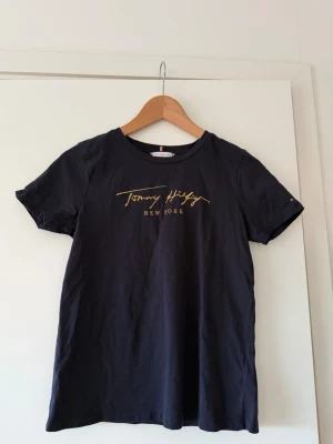 Svart t-shirt Tommy Hilfiger - Svart t-shirt från Tommy Hilfiger med guldfärgat tryck 'Tommy Hilfiger New York' på bröstet. Klassisk rund hals och korta ärmar. 