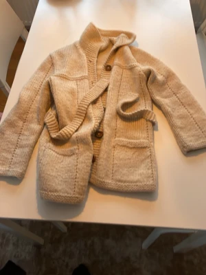 Beige stickad kofta med bälte - Säljer en beige stickad kofta med bälte och stora fickor framtill. Koftan har långärmade ärmar, ribbade kanter och knappar längs framsidan. Perfekt för att hålla sig varm under kyliga dagar.