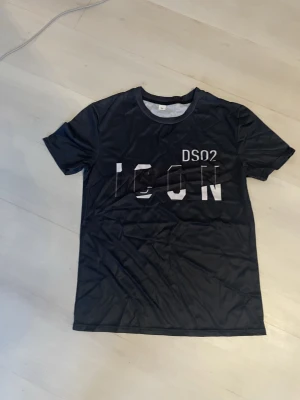 Svart t-shirt med tryck ICON DS02 - Svart t-shirt med vit texttryck 'ICON' och 'DS02' på framsidan. Klassisk rund hals och korta ärmar. Materialet känns lätt och syntetiskt, perfekt för en chill och modern look.