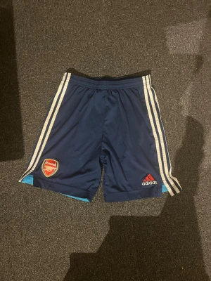 Arsenal x Adidas blå shorts  - Snygga blå Arsenal shorts från Adidas med vita ränder längs sidorna och klubbens emblem på ena benet. Perfekta för fotboll eller träning, med elastisk midja för skön passform. Storlek XL Barn, 176