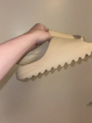 Adidas Yeezy Slide i beige - Säljer ett par Adidas Yeezy Slide i beige färg. Slidesen har en chunky, räfflad sula och är tillverkade i syntetmaterial för extra komfort. De har öppen tå och minimalistisk design med Adidas-logga på fotbädden. Perfekta för chill dagar och sommarhäng.