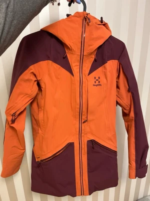 Skidjacka från Haglöfs - Säljer en skidjcka från Haglöfs i storlek woman xs. Använd men i bra skick! Säljer pga för liten.  Den har en huva som är ställbar, en ficka på armen lämpad för skidkort, en bröstficka och två vanliga fickor på magen, samt dragkedjor under armarna för luftning. Jackan är tvåfärgad, orange och vinröd. Skriv om du har någon fråga eller för fler bilder!💕Pris går att diskutera🩷