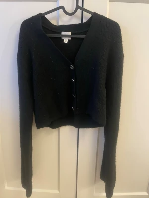 Svart croppad kofta från H&M - Svart croppad kofta från H&M i stickat material. Koftan har långärmade ärmar, v-ringad hals och fyra snygga knappar framtill. Använts fåtal gånger💕