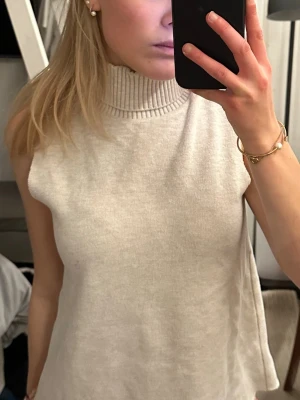 Beige stickad polotröja - Säljer en beige ärmlös stickad polotröja med hög ribbad krage. Tröjan har en loose passform och är perfekt att styla över en skjorta eller långärmad topp. Enkel och stilren design som passar till många outfits. Använd fåtal gånger😊🙌