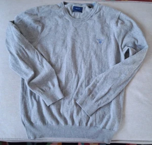 Grå tröja från GANT med broderad logga - Snygg grå tröja från GANT med diskret broderad logga i blått på bröstet. Tröjan har rund hals, långa ärmar och ribbade muddar vid ärmslut och nederkant. Perfekt för dig som gillar klassisk och enkel stil.