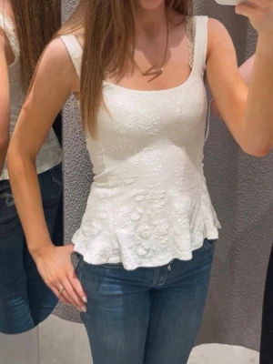 Vit peplum topp med spets  - Säljer ett superfint vitt linne med breda axelband och volangkant nedtill. Toppen har ett blommigt spetsmönster som ger en romantisk vibe och är figurnära i modellen. Perfekt till jeans eller kjol för en fräsch look.