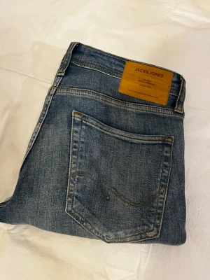 Blå jeans från Jack & Jones - Snygga blå jeans från Jack & Jones med klassisk femficksdesign och kontrastsömmar. Jeansen har en brun läderpatch med logga bak i midjan och är tillverkade i slitstarkt bomullsmaterial. Passar dig som gillar en tidlös och avslappnad look.