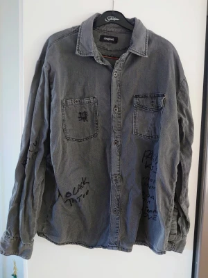 Grå overshirt från Desigual - Säljer en grå overshirt från Desigual med coola handskrivna texter och detaljer på både fram- och baksida. Skjortan har två bröstfickor, knappar hela vägen och lång ärm. Materialet är mjuk bomull och passformen är loose för en avslappnad vibe.