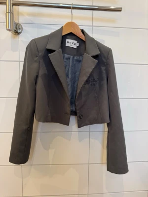 Grå croppad kavaj från NA-KD - Snygg grå croppad kavaj från NA-KD med klassisk krage och långa ärmar. Kavajen har en knapp framtill och fyra dekorativa knappar på ärmsluten. 