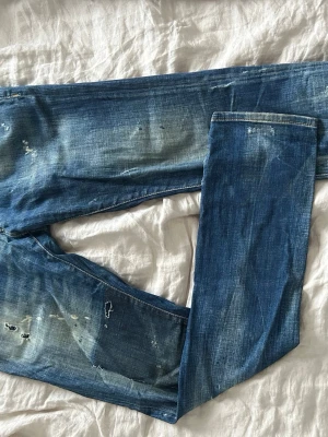 Blå jeans från Dsquared2, storlek 48 - Säljer ett par blå jeans från Dsquared2 med coolt slitet och distressed utseende, vita färgstänk och detaljerad röd Dsquared2-logga baktill. Klassisk femficksmodell med normal passform och midwaist. Storlek 48 och använd ett flertal gånger men har tagit väl hand om dessa. ÄKTA, dessa vill jag bara bli av med 