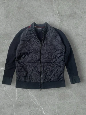 Svart quiltad bomberjacka Tommy Hilfiger | S - Svart bomberjacka från Tommy Hilfiger i storlek L. Framsidan är quiltad med syntetmaterial och har en dragkedja samt en bröstficka med zip. Ärmar och krage är stickade och har en diskret röd detalj vid axeln. Perfekt för dig som gillar stilren och sportig look.