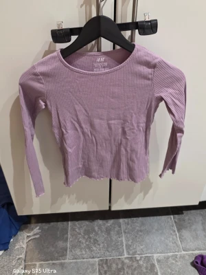Ljuslila ribbad topp från H&M - Säljer en ljuslila långärmad topp från H&M i ribbad bomull med elastan. Toppen har en rund halsringning och vågig kant nedtill för en extra cool touch. Perfekt att matcha med jeans eller kjol för en chill look.