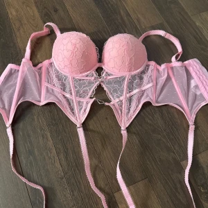 Rosa korsett med hjärtmönster - Säljer en söt rosa korsett med vadderade kupor och hjärtmönster. Den har justerbara axelband och en dekorativ hjärtformad detalj framtill. Baksidan har en rad med hyskor för justering. Perfekt för en romantisk look. Aldrig använd